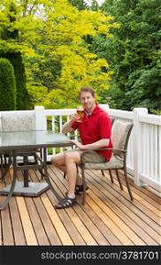 Vertical photo of mature man looking forward and smiling while holding his glass of beer at the table on open cedar patio with seasonal trees in full bloom in background