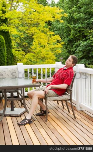 Vertical photo of mature man taking a nap while holding onto his glass of beer while sitting at table on open cedar patio with seasonal trees in full bloom in background