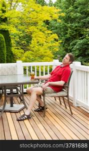 Vertical photo of mature man taking a nap while holding onto his glass of beer while sitting at table on open cedar patio with seasonal trees in full bloom in background