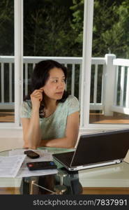 Vertical photo of mature woman thinking while working at home with laptop, cell phone, calculator and papers on top of table and large windows in background