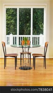 Vertical photo of small dining room with red oak floors, glass table, wooden chairs, bamboo plant and large windows with evergreen trees in background