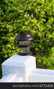 Vertical photo of solar lamp, during day light, on deck post with green trees in background