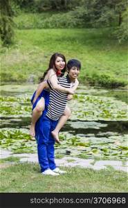 Vertical photo of young adult couple, woman riding on boyfriend back, man holding red rose in mouth with lily pond in background