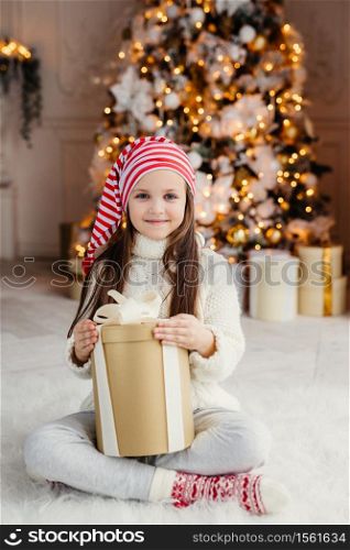 Vertical portrait of beautiful pleasant looking small child wears knitted sweater and socks, sits crossed legs with present, has desire to wrap it, being in living room near decorated New Year tree