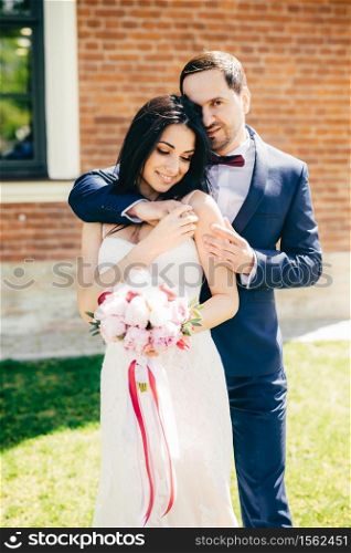Vertical portrait of brunette beautiful charming young bride in white dress, recieve embrace from affectionate bridegroom, have good relationship and great love to each other. Wedding concept