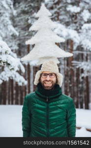 Vertical portrait of cheerful bearded man has fun alone in winter forest, keeps artificial fir tree, poses outdoors, admires frosty snowy weather, expresses positiveness and pleasant emotions