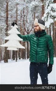 Vertical portrait of dlightful positive bearded male dressed in warm winter clothes, looks with happy expression at artificial fir tree, demonstrates that he protects nature and doesn`t cut down trees