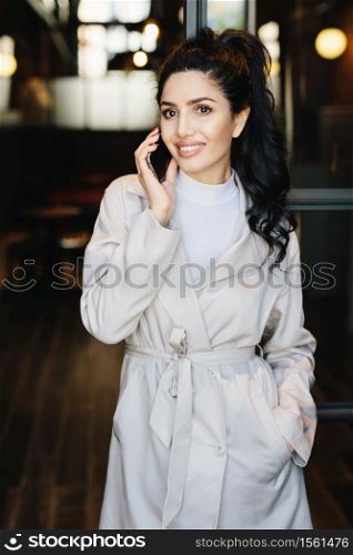 Vertical portrait of elegant brunette businesswoman in white coat communicating over cell phone while standing outdoors. Caucasian woman with beautiful appearance using modern gadget. Fashion concept