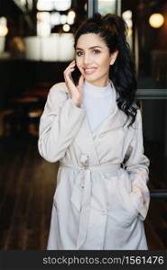 Vertical portrait of elegant brunette businesswoman in white coat communicating over cell phone while standing outdoors. Caucasian woman with beautiful appearance using modern gadget. Fashion concept