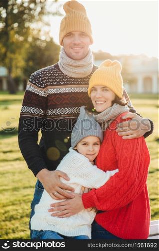 Vertical portrait of friendly family couple stand together, embrace each other, have good relationships, enjoy sunny weather. Handsome unshaven man hugs his wife and daughter, pose outdoors