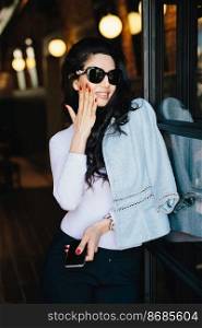 Vertical portrait of glamour businesswoman with dark luxurious hair wearing sunglasses and formal clothes holding hand on cheek demonstrating her red nice nails using cell phone while standing in cafe