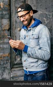 Vertical portrait of hipster guy in denim shirt, cap and eyewear holding modern telephone in his hands having glad expression while communicating with his girlfriend. Lifestyle and technolgy concept