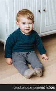Vertical portrait of infant sits on floor near cupboard, looks with warm blue eyes. Indoor shot of handsome blonde baby male going to play with children. Small toddler look curiously into camera