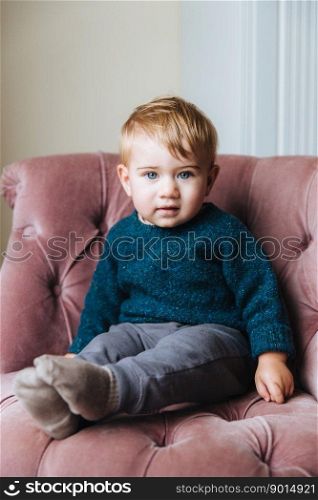 Vertical portrait of innocent little child with blue eyeys and plump cheeks, looks directly into camera, wait for children as has birthday, sits in comfortable pink armchair in living room. Children