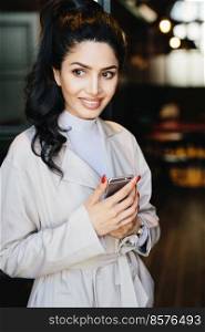 Vertical portrait of lovely brunette woman in stylish clothes having red manicure holding smartphone in hands having pensive look aside. Attractive model with dark eyes and smile looking aside