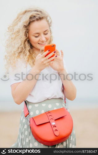 Vertical portrait of lovely young woman has light curly hair, holds modern cellular, happy to see photo of lover in social networks, carries small bag, stands outdoor at coastline. Lifestyle concept