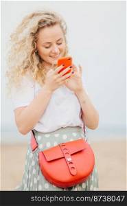 Vertical portrait of lovely young woman has light curly hair, holds modern cellular, happy to see photo of lover in social networks, carries small bag, stands outdoor at coastline. Lifestyle concept