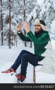 Vertical portrait of pleasant looking male makes photo with smart phone, makes shots of beautiful winter landscapes, enjoys nature and frozy weather, has pleasant smile. People, technology concept