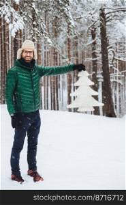 Vertical portrait of positive smiling male wears fashionable warm clothes, keeps white fir tree in hands, poses in white winter forest. Happy unshaven attractive man spend free time or winter holidays