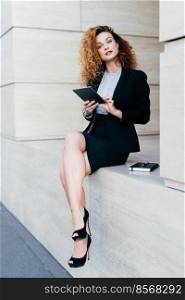 Vertical portrait of pretty slim woman with curly hair, wearing black jacket, skirt and high-heeled shoes, using modern tablet computer for searching information for her work, writing in notebook