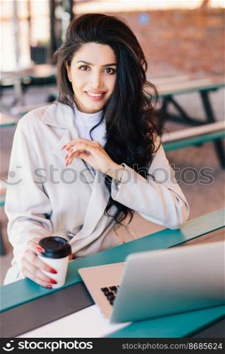 Vertical portrait of young brunette female entrepreneur enjoying rest at cafe with electronic device and coffee checking email on laptop, using free wireless connection looking directly into camera