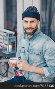 Vertical shot of attractive blue eyed bearded male in stylish clothing, drinks hot tea, looks with pleased expression at camera, spends recreation time at terrace cafe, smiles gently at camera