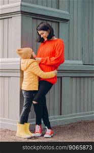 Vertical shot of attractive caring mother looks at eyes of her daughter with love, embrace while pose outside, wears ripped jeans, sweater and sneakers, smile positively. Family relationship concept