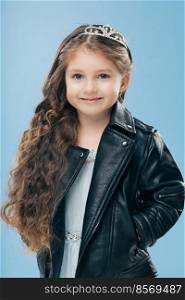 Vertical shot of attractive small female child has long curly hair, wears crown and leather jacket smiles positively isolated over blue wall. Happy little girl dressed in stylish clothes models indoor