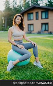 Vertical shot of beautiful brunette woman sits on fitness ball over green grass and house in background, dressed in active wear, has yoga or gymnastic training, being in good shape. Active lifestyle