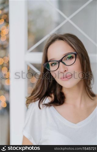 Vertical shot of charming woman in optical glasses, dressed casually, poses indoor against Christams tree, has healthy soft skin, enjoys spare time in domestic atmosphere. People and leisure