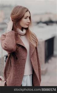 Vertical shot of fashionable Caucasain woman looks aside, wears fashionable warm coat, poses outdoor, enjoys day off, has date. Beautifu young lady goes to work, carries womans bag. Urban style