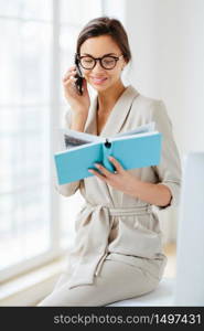 Vertical shot of female administrative manager involved in working process, holds notepad with written notes, discusses something via cellphone, wears spectacles and elegant costume, smiles gently