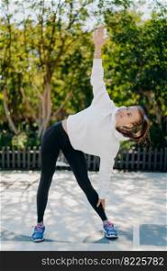Vertical shot of glad sportswoman with combed pony tail≤ans aside keeps o≠arm raised up does exercises outdoors wears white hoodie≤ggings trai≠rs relaxes on nature has active fit≠ss training