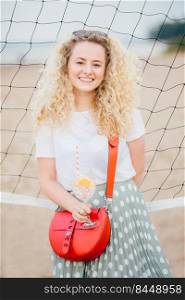 Vertical shot of good looking curly female wears t shirt and skirt, has small bag, holds cocktail with ice, stands near tennis net on beach, has good rest. Lovely woman with appealing charming smile
