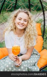 Vertical shot of good looking pretty young blonde female with curly hair, positive smile, spends free time with friends, have party together, poses in hanging chair, drinks fresh summer cocktail.