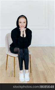 Vertical shot of good looking woman with make up, keeps hands under chin, wears hoody, leggings and white sportshoes, has slender legs, poses on chair in empty room, ready for sport training
