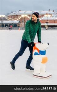 Vertical shot of handsome bearded male being on ice rink, learns to skate with aid or prepares for skating competitions, has happy exression, smiles joyfully. People, winter, hobby concept