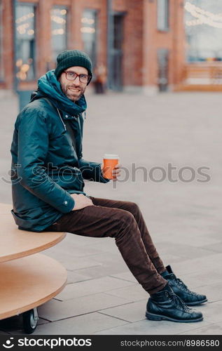 Vertical shot of handsome bearded man dressed in street apparel, drink coffee to go, poses in urban setting, enjoys good rest, being in good mood. Street style and rest concept. Hipster with drink