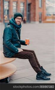 Vertical shot of handsome bearded man dressed in street apparel, drink coffee to go, poses in urban setting, enjoys good rest, being in good mood. Street style and rest concept. Hipster with drink