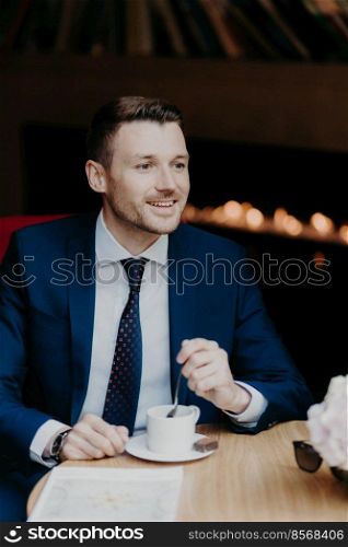 Vertical shot of handsome male in formal clothing, has cheerful expression, has dinner break at cafeteria, drinks aromatic coffee, looks happily somewhere, thinks about something. Business and rest
