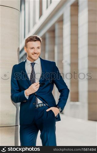 Vertical shot of handsome male with appealing appearance, dressed in formal black suit, keeps hand in pocket, waits for business partners, going to have negotiations, has positive expression.