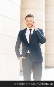 Vertical shot of handsome successful male financial director solves problems via smart phone, looks confidently at camera, keeps hand in pocket, poses outdoor. People and technology concept.