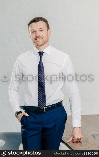 Vertical shot of handsome unshaven cheerful man wears formal clothes, keeps hand in pocket, stand near office table, has positive smile. Prosperous businessman being at work place. Business concept