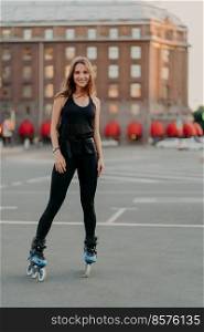 Vertical shot of healthy slim woman going rollerblading dressed in active wear spends free time outdoors smiles happily has exercising activities leads sporty lifestyle. Inline skating concept