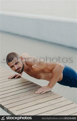 Vertical shot of muscular bearded man stands in plank pose trains chest and arms muscles has hard workout for keeping athletic body shape poses outdoor. Active lifestyle and motivation concept