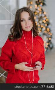 Vertical shot of pleasant looking brunette female model dressed in loose warm sweater, carries smart phone, uses earphones for listening music, poses in domestic interior near decorated fir tree