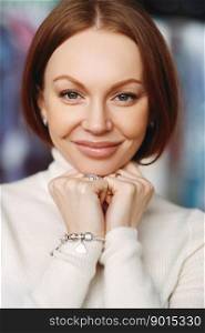Vertical shot of pleasant looking lady with brown hair, keeps hands under chin, has healthy skin, make up, wears bracelet and white jumper, looks at camera, has broad forehead, nice appearance