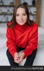 Vertical shot of pleasant looking woman in casual red knitted sweater, has manicure, enjoys domestic atmosphere, has make up, poses indoor. Beautiful female with dark hair spends free time at home