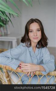 Vertical shot of pleasant looking young European dark haired woman sits on wicker chair, dressed in striped jacket, has red manicure, has charming look at camera, poses against domestic interior.