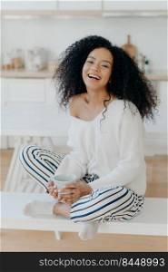 Vertical shot of pleased young Afro American woman feels relaxed and carefree, drinks aromatic coffee, sits crossed legs against kitchen interior, being in good mood, spends spare time at home